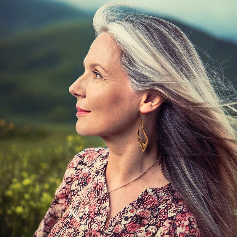 A head shot of a woman in a field looking to the left with a laser cut wooden earring in the shape of a feather hanging off her head, her hair blowing back slightly, she is wearing a flower dress.